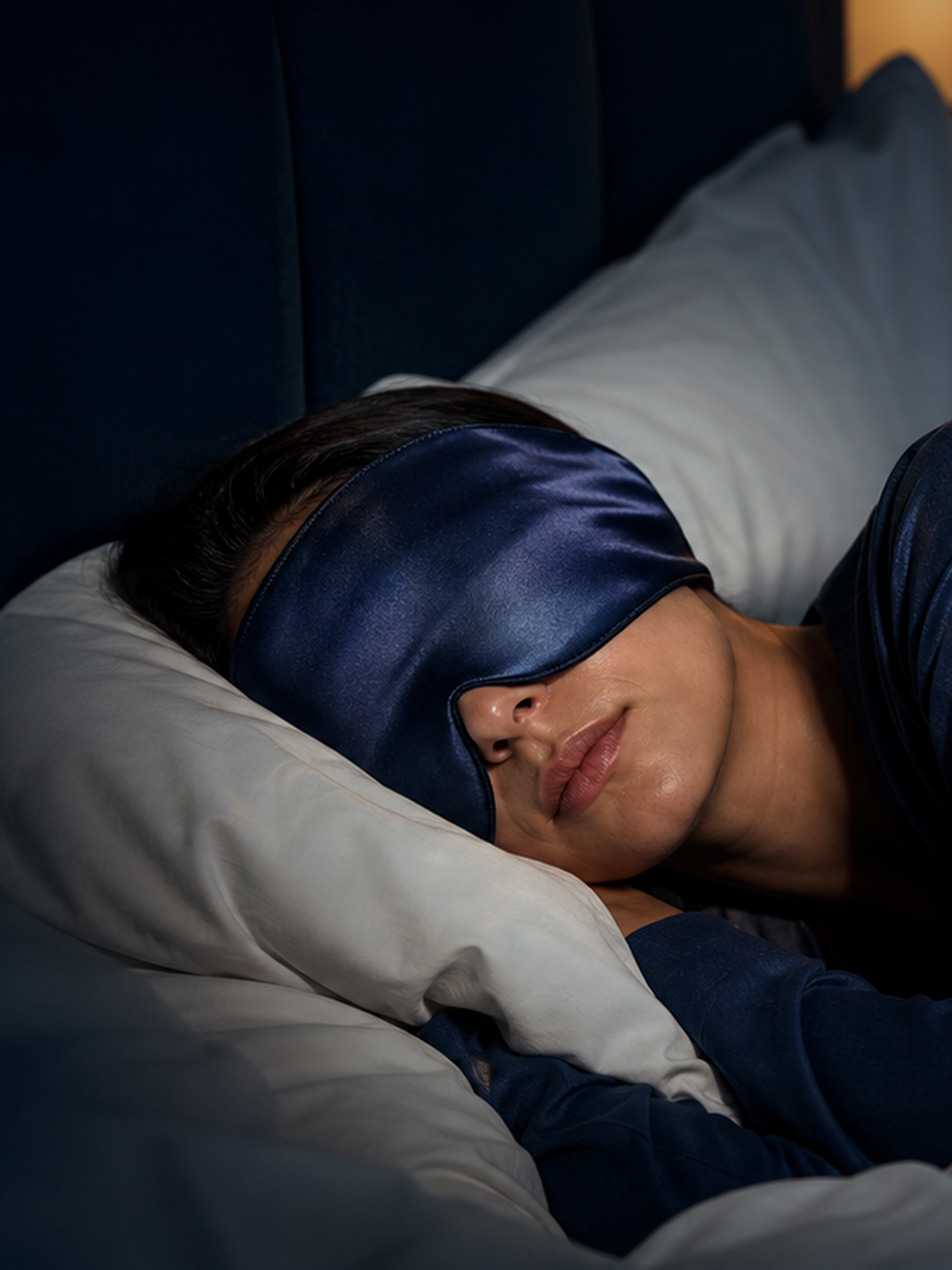 A person lying in bed wearing a dark blue satin sleep mask and dark blue pajamas, resting on white pillows against a dark headboard.